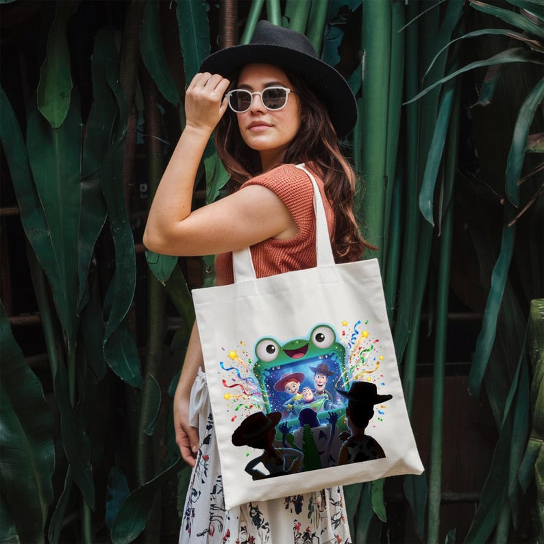 May include: A white canvas tote bag featuring a colorful graphic of a green frog-shaped television screen displaying animated characters, surrounded by confetti. The bag has two sturdy shoulder straps and is made of a durable fabric material.
