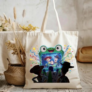 May include: A cream-colored canvas tote bag featuring a graphic print of a green frog-shaped television screen displaying characters from Toy Story. The design includes colorful confetti and silhouettes of characters watching the screen.