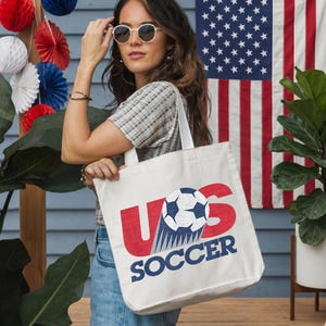 Pode incluir: Uma sacola de lona branca com um design gr&aacute;fico contendo as letras vermelhas US, uma bola de futebol preta e branca substituindo a letra O e a palavra SOCCER impressa em azul abaixo. O design tem uma apar&ecirc;ncia texturizada e desgastada.