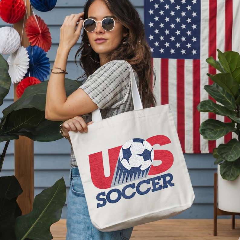 Pode incluir: Uma sacola de lona branca com um design gr&aacute;fico contendo as letras vermelhas US, uma bola de futebol preta e branca substituindo a letra O e a palavra SOCCER impressa em azul abaixo. O design tem uma apar&ecirc;ncia texturizada e desgastada.