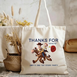 May include: A cream-coloured canvas tote bag featuring a graphic of Woody and Jessie riding Bullseye from Toy Story. The text reads "THANKS FOR racing by!" at the top and "AMY'S 2ND TOY STORY PARTY" at the bottom in dark blue lettering.
