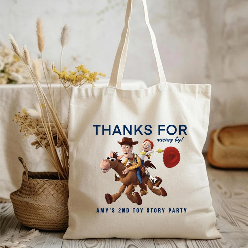 May include: A cream-coloured canvas tote bag featuring a graphic of Woody and Jessie riding Bullseye from Toy Story. The text reads "THANKS FOR racing by!" at the top and "AMY'S 2ND TOY STORY PARTY" at the bottom in dark blue lettering.