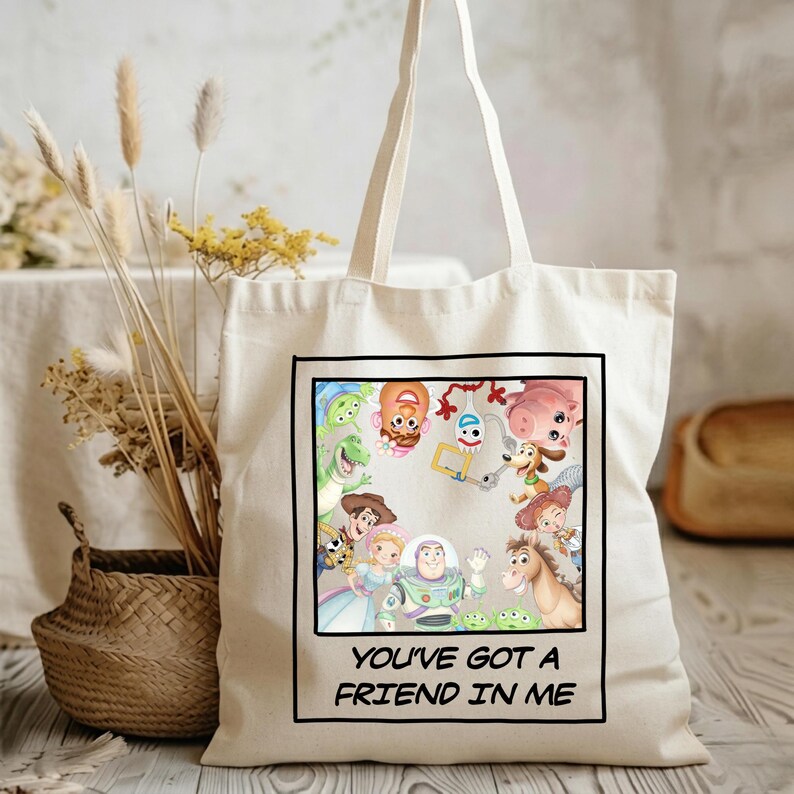 May include: A cream-colored canvas tote bag featuring a square graphic print of various Toy Story characters arranged in a circle. Below the illustration, the text reads "YOU'VE GOT A FRIEND IN ME" in black capital letters within a black border.