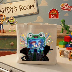 May include: A cream-colored canvas tote bag featuring a colorful graphic print of a green frog-shaped television screen displaying characters from the Toy Story franchise, surrounded by confetti, with silhouettes of two figures in the foreground.