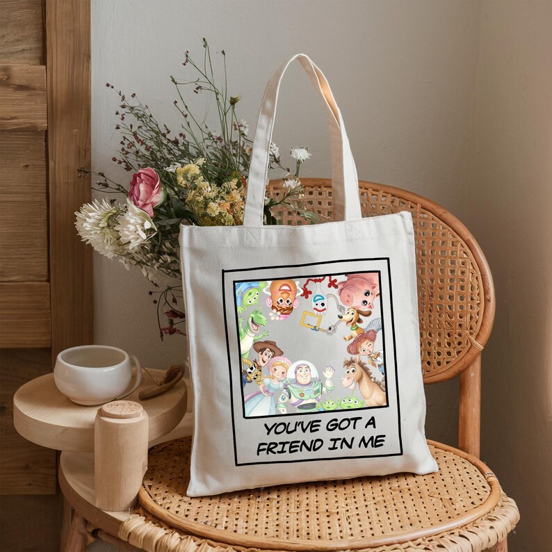 May include: A white canvas tote bag featuring a square graphic print of various animated characters arranged in a circle. Below the graphic, the text reads YOU'VE GOT A FRIEND IN ME in black capital letters. The bag has two matching fabric handles.