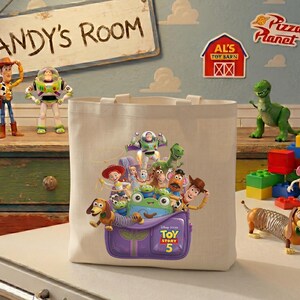 May include: A cream-colored canvas tote bag featuring a colorful graphic of Toy Story characters emerging from a purple backpack. The design includes Woody, Buzz Lightyear, Jessie, Slinky Dog, and Mr. Potato Head, with the text Toy Story 5 printed below.