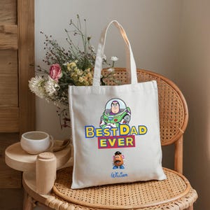 May include: A white canvas tote bag featuring a graphic of Buzz Lightyear above the text BEST DAD EVER in blue and yellow letters, with a Mr. Potato Head character below and the name William written in blue script at the bottom of the bag.