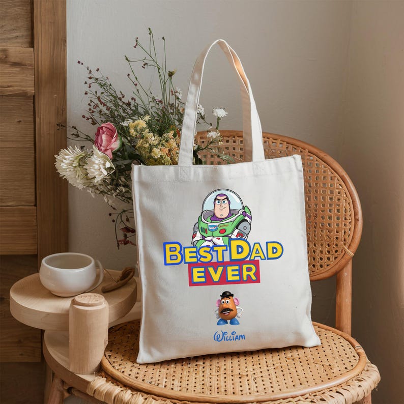 May include: A white canvas tote bag featuring a graphic of Buzz Lightyear above the text BEST DAD EVER in blue and yellow letters, with a Mr. Potato Head character below and the name William written in blue script at the bottom of the bag.