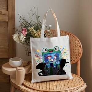 May include: A white canvas tote bag featuring a colorful graphic print of a green frog-shaped television screen displaying characters from Toy Story, surrounded by festive confetti and streamers, with silhouettes of characters in the foreground.