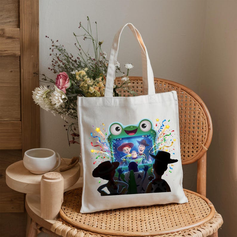 May include: A white canvas tote bag featuring a colorful graphic print of a green frog-shaped television screen displaying characters from Toy Story, surrounded by festive confetti and streamers, with silhouettes of characters in the foreground.