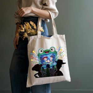 May include: A cream-colored canvas tote bag featuring a graphic print of a green frog-shaped television screen. Inside the screen, characters from Toy Story are visible. The design includes colorful confetti and streamers surrounding the television.