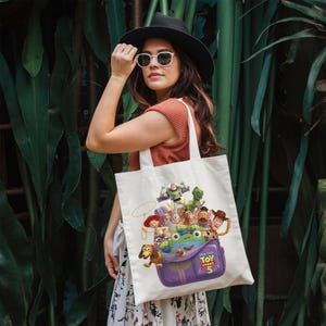 May include: A white canvas tote bag featuring a colorful graphic print of Toy Story 5 characters, including Woody, Buzz Lightyear, Jessie, and Slinky Dog, emerging from a purple bag. The design includes the Toy Story 5 logo in yellow and red text.