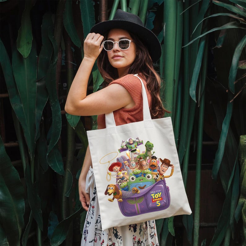 May include: A white canvas tote bag featuring a colorful graphic print of Toy Story 5 characters, including Woody, Buzz Lightyear, Jessie, and Slinky Dog, emerging from a purple bag. The design includes the Toy Story 5 logo in yellow and red text.