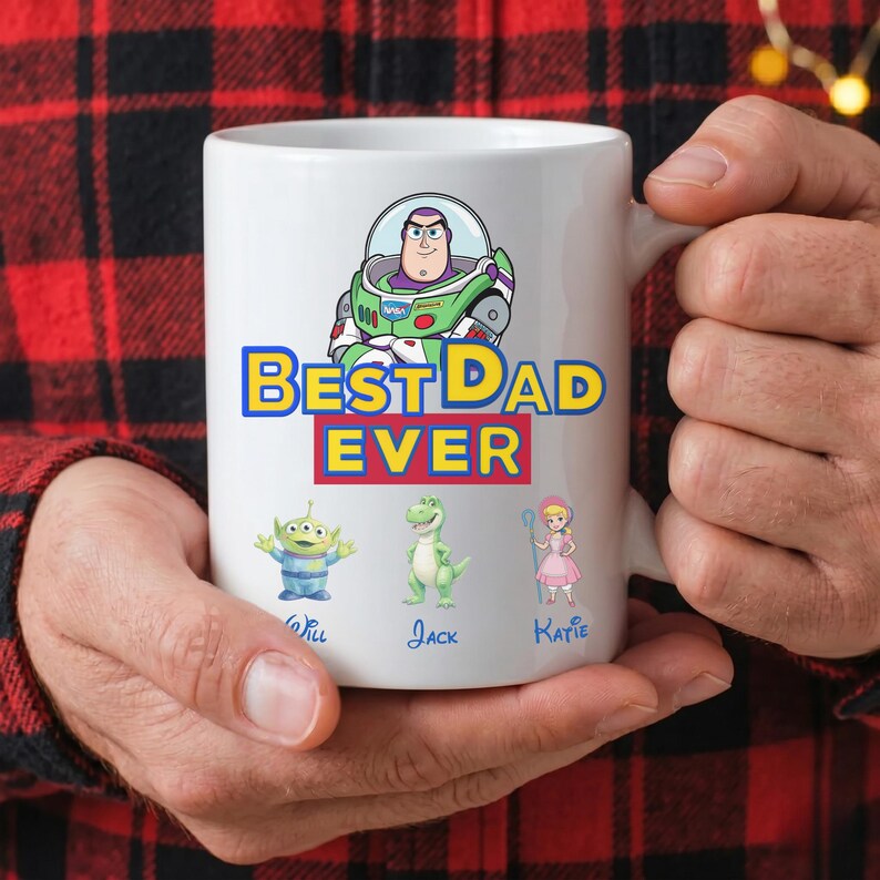 Puede incluir: Una taza de cer&aacute;mica blanca con un gr&aacute;fico de Buzz Lightyear en la parte superior, el texto BEST DAD EVER en azul y rojo, y tres personajes debajo etiquetados como Will, Jack y Katie, incluyendo un alien&iacute;gena, un dinosaurio verde y una mu&ntilde;eca rubia.