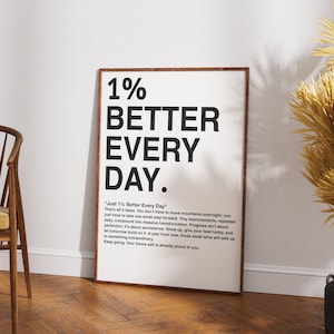 May include: A framed poster with the text "1% BETTER EVERY DAY" in bold black letters. The poster has a wooden frame and is leaning against a white wall. A wooden chair and decorative plants are also in the image.