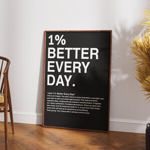 May include: A framed black and white print with the text "1% BETTER EVERY DAY." The print has a wooden frame and includes additional text below the main title. The print is leaning against a white wall.