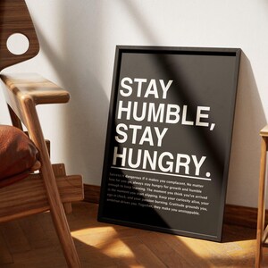 Op de afbeelding: Zwartgelijste poster met witte tekst "STAY HUMBLE, STAY HUNGRY". De poster staat tegen een witte muur, naast een houten stoel met een bruine leren zitting. Er staat ook een alinea tekst onder de hoofdtekst.