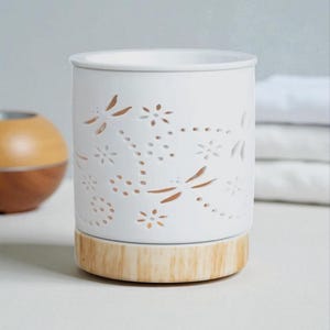 May include: White ceramic essential oil diffuser with cut-out dragonfly and floral designs. The diffuser has a light wood-colored base and a cylindrical shape. The design is a decorative home fragrance accessory.