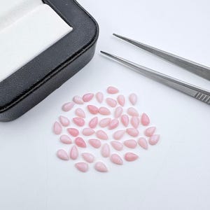 Peut inclure: Une collection de petits cabochons rose clair en forme de poire disposés en grappe. Une paire de pinces en métal argenté repose à proximité, et une boîte de présentation à bijoux noire avec un intérieur blanc est placée dans le coin supérieur gauche.