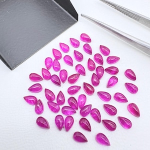 May include: A collection of small, pear-shaped, vibrant pink gemstones arranged on a white surface. A black metal scoop and a pair of silver metal tweezers are positioned near the loose stones, which feature a faceted cut and a uniform color.