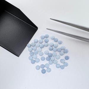 May include: A collection of small, round, light blue cabochon gemstones arranged in a cluster. A black metal scoop is positioned to the left, and a pair of silver metal tweezers is placed to the right of the stones on a plain white surface.