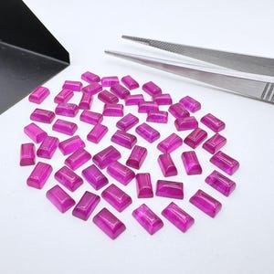 Puede incluir: Una colección de pequeñas piedras preciosas rectangulares de color magenta con una superficie superior lisa y abovedada. Las piedras están dispuestas en un grupo suelto junto a unas pinzas de metal plateado y una pala de metal negro.