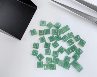 May include: A collection of small, square-cut green aventurine gemstones with a faceted pyramid top. The stones are scattered near a black metal scoop and a pair of silver metal tweezers, showcasing their translucent, light green color and smooth, polished finish.
