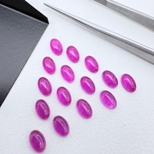 Peut inclure: Un ensemble de treize petites pierres précieuses ovales d'un magenta vif disposées en triangle. Une paire de pinces en métal argenté est placée en haut à droite, et un coin de plateau en métal noir est visible sur le côté gauche.