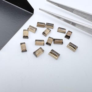 Könnte beinhalten: Eine Ansammlung kleiner, rechteckiger, durchscheinender Rauchquarz-Edelsteine mit facettierter Oberfläche. Die Steine sind neben einer schwarzen Metallschaufel und einer silbernen Metallpinzette angeordnet und zeigen ihre braune, halbtransparente Optik.