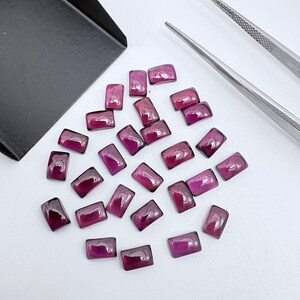 Pode incluir: Uma coleção de pequenas pedras preciosas retangulares de cor magenta profundo com superfícies lisas e polidas. As pedras estão dispostas num grupo solto ao lado de uma pá de metal preta e uma pinça de metal prateado sobre um fundo branco.