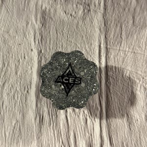 May include: A silver glittery, scalloped-edge circular object featuring a black diamond-shaped logo with the text ACES printed in the center. The surface is textured with embedded reflective particles that catch the light across the entire piece.