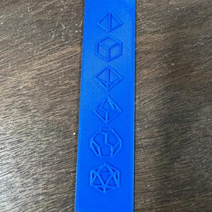 May include: A vibrant blue, rectangular game piece featuring embossed geometric shapes of various dice. The shapes include a tetrahedron, cube, octahedron, and a dodecahedron, and an icosahedron. The piece is likely for a tabletop role-playing game.
