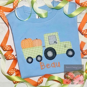 May include: A light blue t-shirt with an embroidered design of a green tractor pulling a wagon full of orange pumpkins. The name "Beau" is embroidered below the tractor.