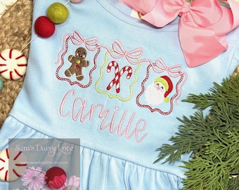May include: A light blue Christmas-themed dress with embroidered gingerbread man, candy canes, and Santa Claus designs. The name "Camille" is embroidered below the designs. A large pink bow and decorative garland are also visible.