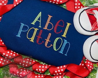 May include: A navy blue baby onesie with the name "Abel Patton" embroidered in rainbow colors. The onesie is surrounded by red and white polka dot ribbon.