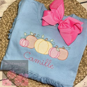 May include: A light blue long-sleeved shirt with a ruffle hem and a large pink bow. The shirt features an embroidered design of pumpkins in shades of pink and yellow, with the name "Camille" stitched below.