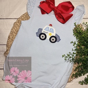 May include: Light blue and white striped romper with a police car applique. The police car has a white body, blue and white checkered accents, and a yellow star. A large red bow is at the top. The romper has ruffle sleeves.