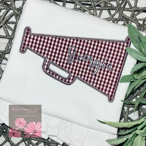 May include: White t-shirt featuring a red and white gingham megaphone design. The megaphone has the word "Georgia" embroidered in silver thread.