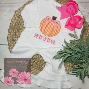May include: A white long-sleeve romper with ruffled cuffs and hem, featuring an orange gingham pumpkin appliqué with a pink bow and the name Margaret embroidered in pink. A large bright pink hair bow is attached to the shoulder area.