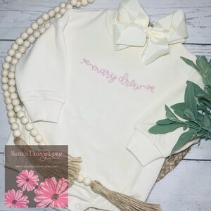 May include: White long-sleeve baby onesie with pink embroidered text "mary drew" on the front.