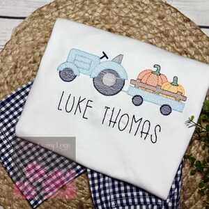 May include: White t-shirt embroidered with a light blue tractor pulling a wagon with two pumpkins. The name "LUKE THOMAS" is embroidered below the tractor. The shirt is on a blue and white checkered fabric.