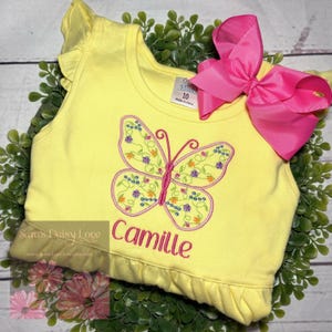 May include: A yellow top with a butterfly design embroidered with pink and floral accents. The name "Camille" is embroidered in pink below the butterfly. A large pink bow is attached. The top has a ruffled hem and sleeve detail.