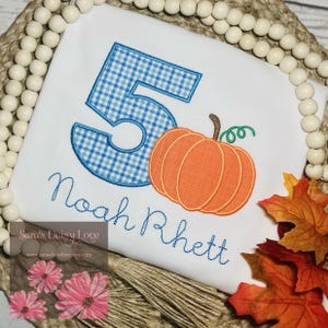 May include: A white onesie with a blue and white gingham number 5, an orange pumpkin, and the name "Noah Rhett" embroidered in white thread.