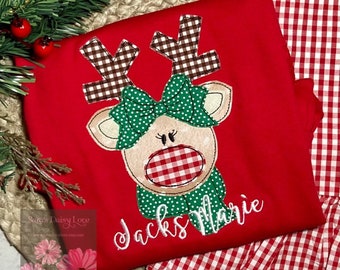 May include: A red shirt featuring an applique of a reindeer with brown and white gingham antlers, a green polka dot bow, a red and white gingham nose, and a green polka dot scarf. The name Jacks Marie is embroidered in white cursive script.