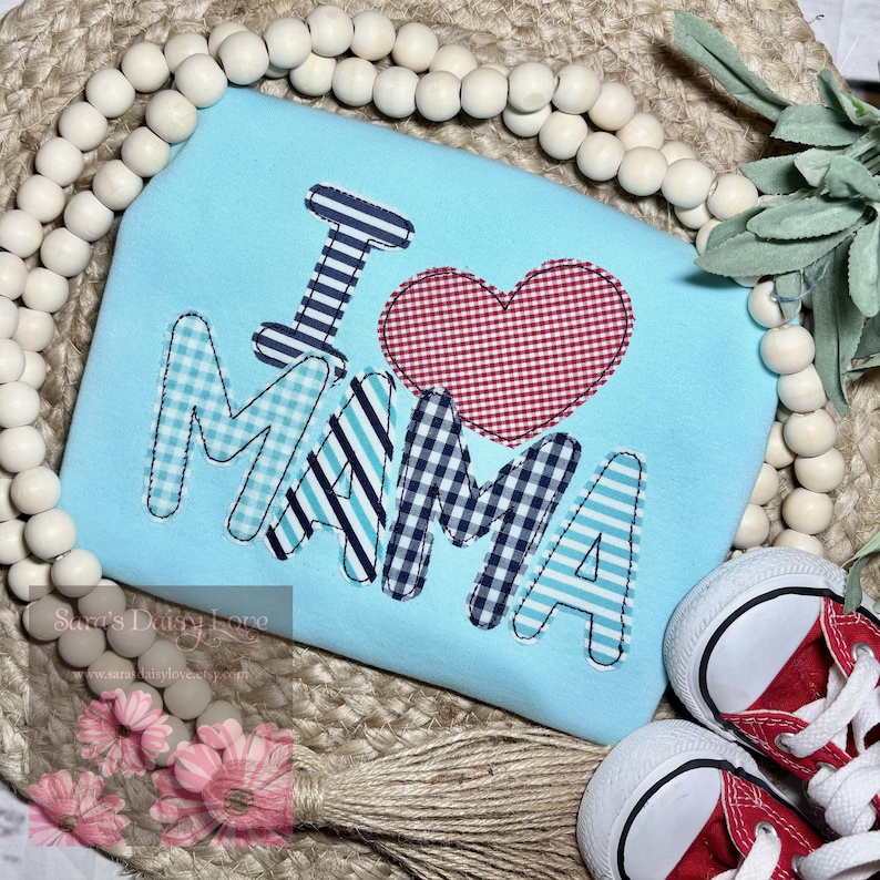 May include: A light blue shirt featuring the words "I love Mama" in colourful patterned fabric. The letters are outlined in white and the "love" is represented by a red and white gingham heart.