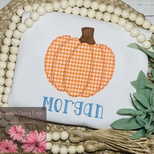 May include: White onesie with a gingham pumpkin and the name "Morgan" embroidered in blue thread. The onesie is on a background of white beads and green foliage.