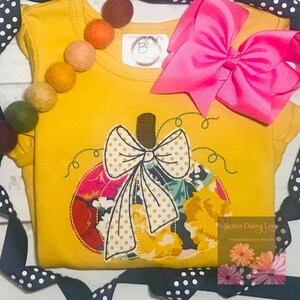 May include: Mustard yellow short-sleeved shirt with a pumpkin applique. The pumpkin is made of floral fabric and has a white polka dot bow. A large pink bow and felt balls are also in the image. The text "Sara's Daisy Love" is visible.