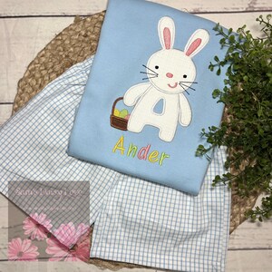 May include: A light blue Easter-themed outfit featuring a bunny design with a basket of eggs and the name "Ander" embroidered in colorful thread. The outfit includes a top and checkered shorts, perfect for the holiday.