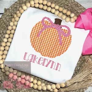 May include: White t-shirt featuring an orange and white gingham pumpkin applique with a pink bow and brown stem. The name "Lakelynn" is embroidered in pink script below the pumpkin. A fall-themed design.
