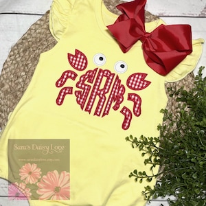 May include: A yellow baby bodysuit with a crab design. The crab features a red and white gingham pattern and the initials "SRM". A large red bow is attached. The bodysuit has ruffled sleeves.